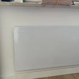 Biały panel grzewczy UDEN-S zamontowany na ścianie pod oknem, widoczny fragment drewnianej podłogi i listwy przypodłogowej.