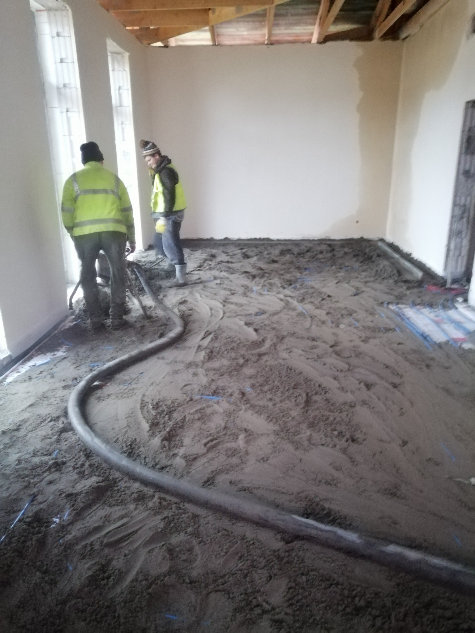 Dwóch pracowników w odblaskowych kamizelkach rozprowadza szary materiał na podłodze, widoczny wąż podający materiał i ogrzewanie podłogowe.