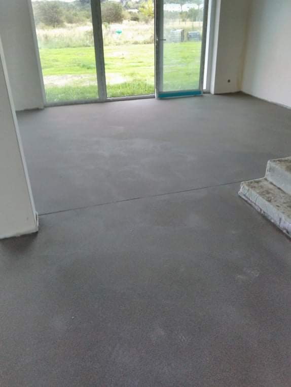 Świeżo wylana, szara posadzka w jasnym pomieszczeniu z dużymi oknami wychodzącymi na zielony teren. Widoczne niedoskonałości powierzchni i fragment schodów.