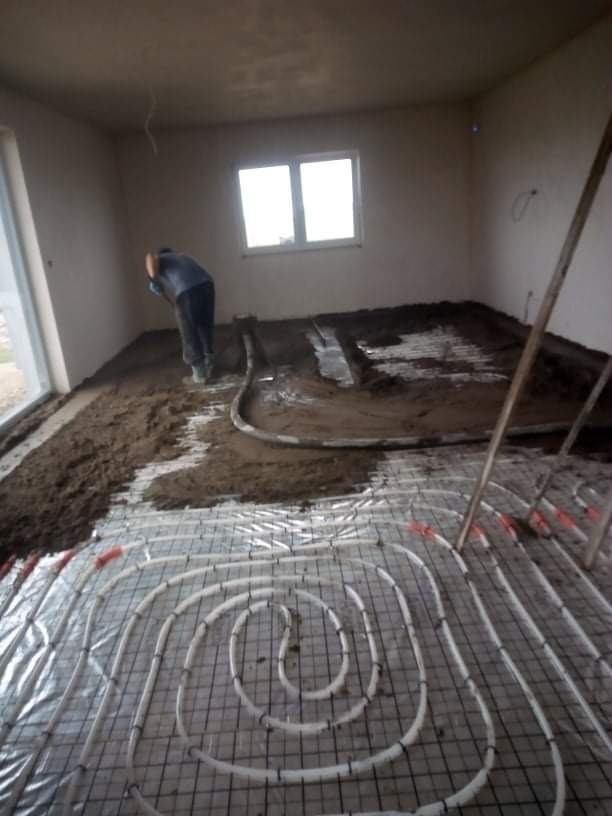 Prace wewnątrz budynku: układanie ogrzewania podłogowego z rur na folii aluminiowej, częściowo zalane masą wyrównawczą, widoczny pracownik w tle.