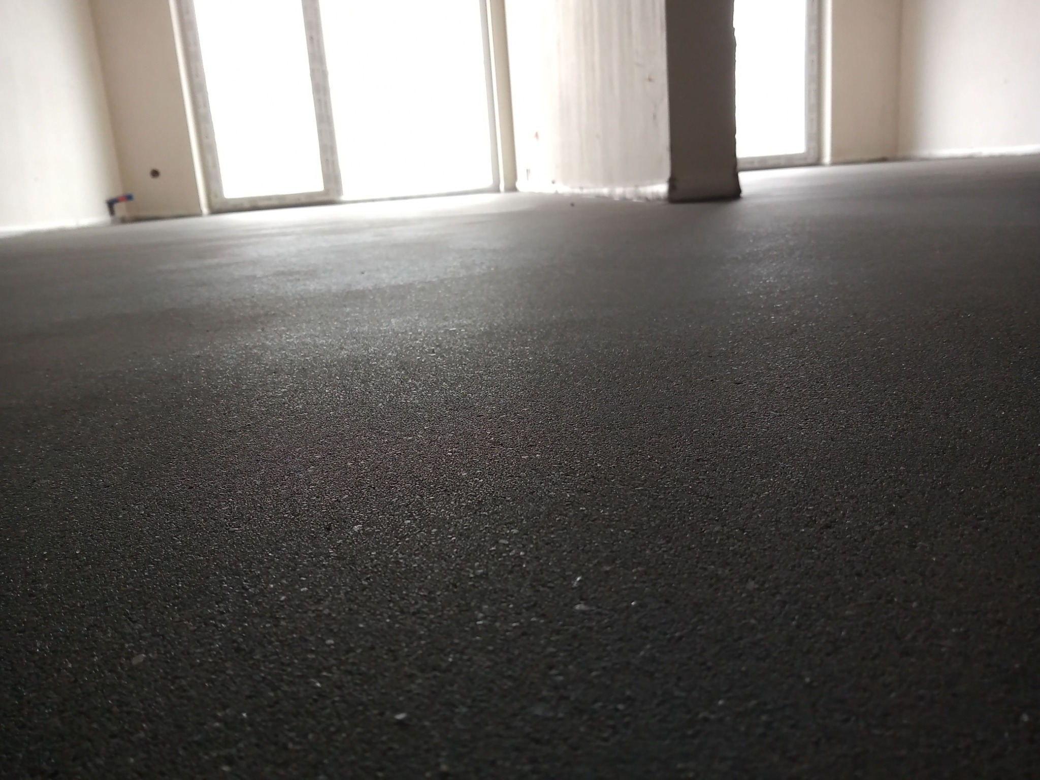 Świeżo wylana, ciemna posadzka w surowym wnętrzu z oknami i słupem konstrukcyjnym, widok z niskiej perspektywy.