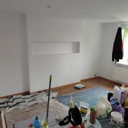 Pokój w trakcie odświeżania, ściany świeżo pomalowane na biało, zabezpieczona folią podłoga, widoczne narzędzia malarskie, puszki z farbą i napoje na szklanym stoliku.
