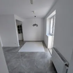 Wnętrze pokoju po remoncie z szarymi płytkami imitującymi marmur na podłodze, białymi ścianami, grzejnikiem i nowoczesną lampą wiszącą.