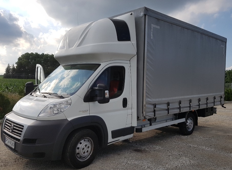 Biały samochód dostawczy z plandeką, otwarte drzwi kierowcy, zaparkowany na żwirowym podłożu z widokiem na pole i drzewa w tle.