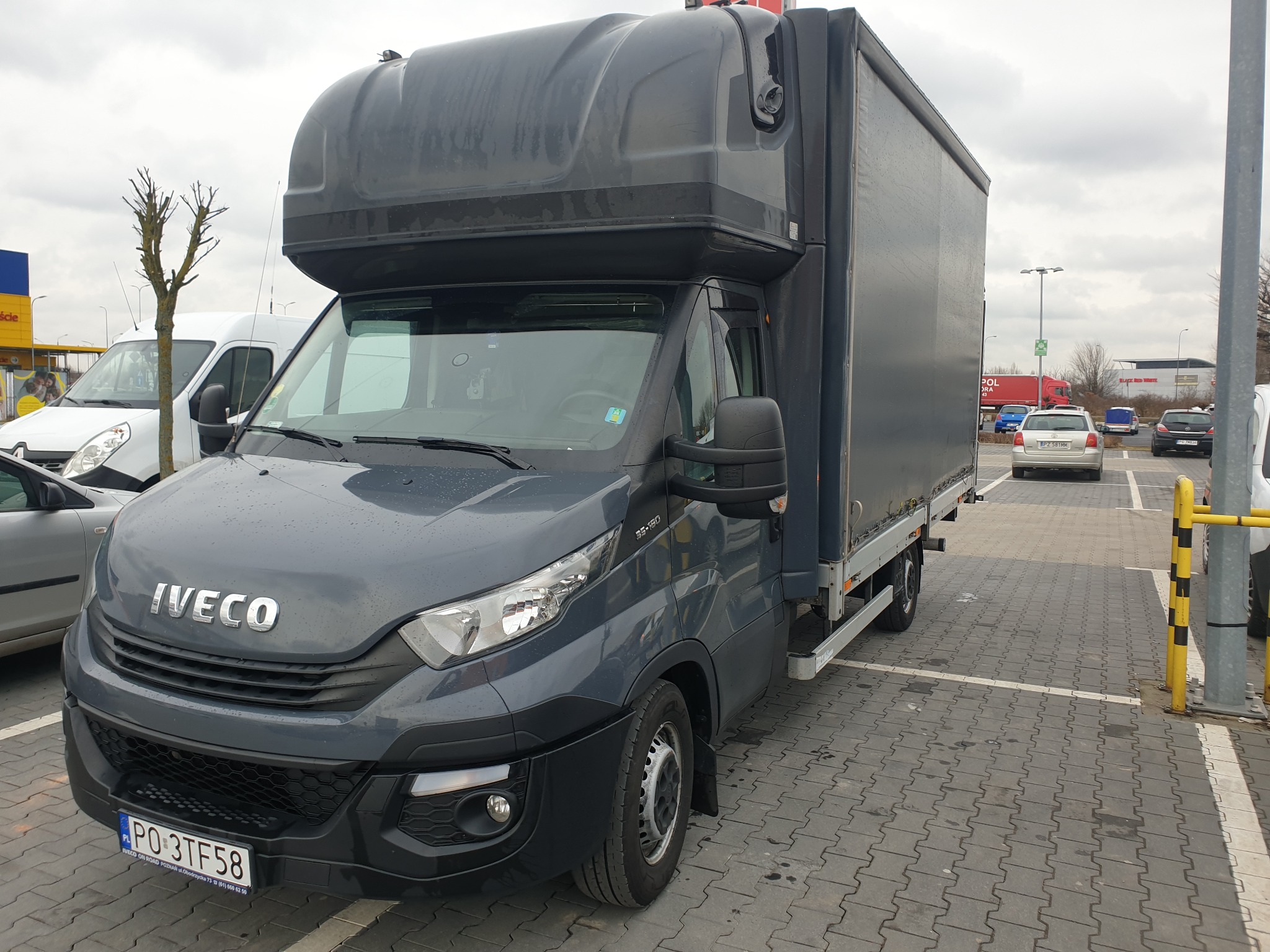 Szary samochód dostawczy marki Iveco z wysoką zabudową, zaparkowany na parkingu z kostki brukowej w pochmurny dzień.