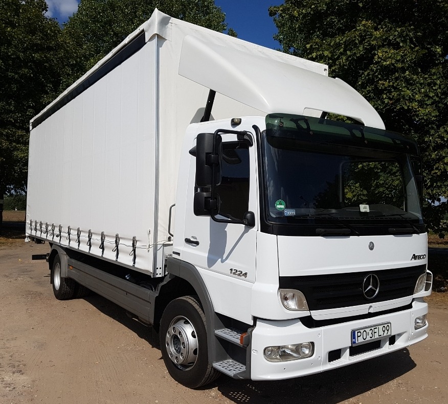 Biały samochód ciężarowy Mercedes Atego 1224 z plandeką, zaparkowany na ziemnej powierzchni w słoneczny dzień.