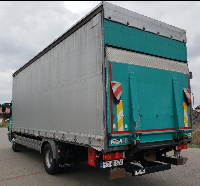 Tylna część ciężarówki z plandeką w kolorze szarym i zielonym, widoczne tablice ostrzegawcze i rejestracyjna, hydrauliczny podnośnik załadowczy.