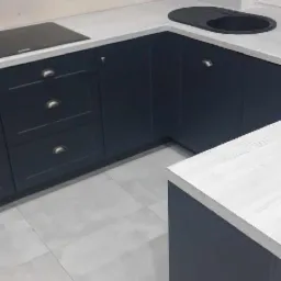 Ciemnogranatowa zabudowa kuchenna z białym blatem, widoczny zlew i płyta indukcyjna, szare płytki podłogowe.