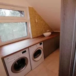 Pralka i suszarka pod blatem drewnianym w łazience na poddaszu ze skośnym sufitem i żółtą mozaiką na ścianie.