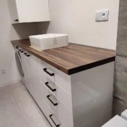 Biała, błyszcząca szafka łazienkowa z drewnianym blatem i prostokątną umywalką nablatową, czarne uchwyty, pralka w zabudowie obok, szara płytka podłogowa.