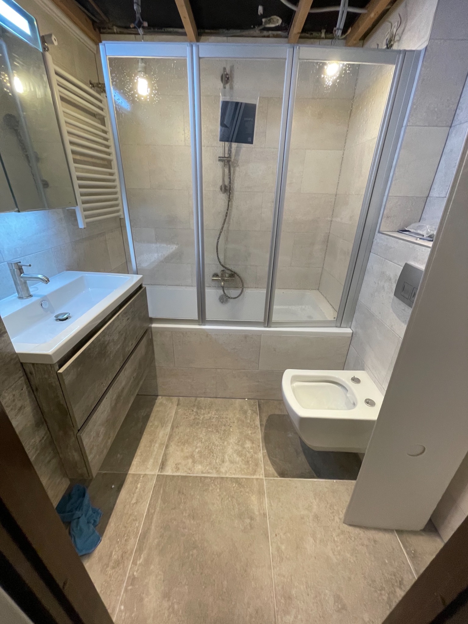 Wnętrze łazienki w trakcie wykańczania, widoczna kabina prysznicowa z wanną, umywalka z szafką w odcieniach szarości, bidet oraz fragment surowego sufitu z widocznymi instalacjami.