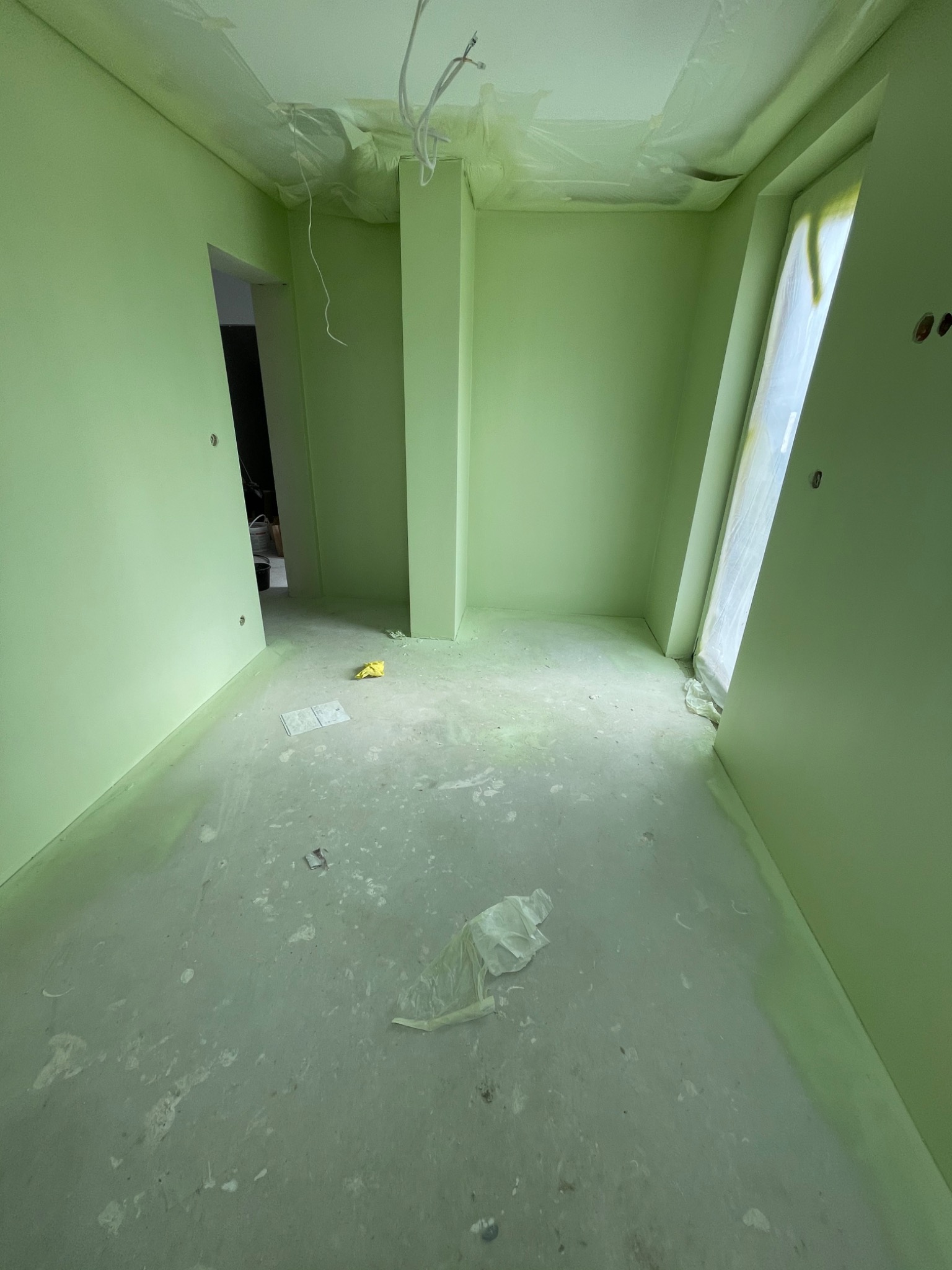 Wnętrze w trakcie malowania na pistacjowy kolor, z widocznymi zabezpieczeniami folią malarską na oknie i przewodach sufitowych, oraz zabrudzeniami na podłodze.