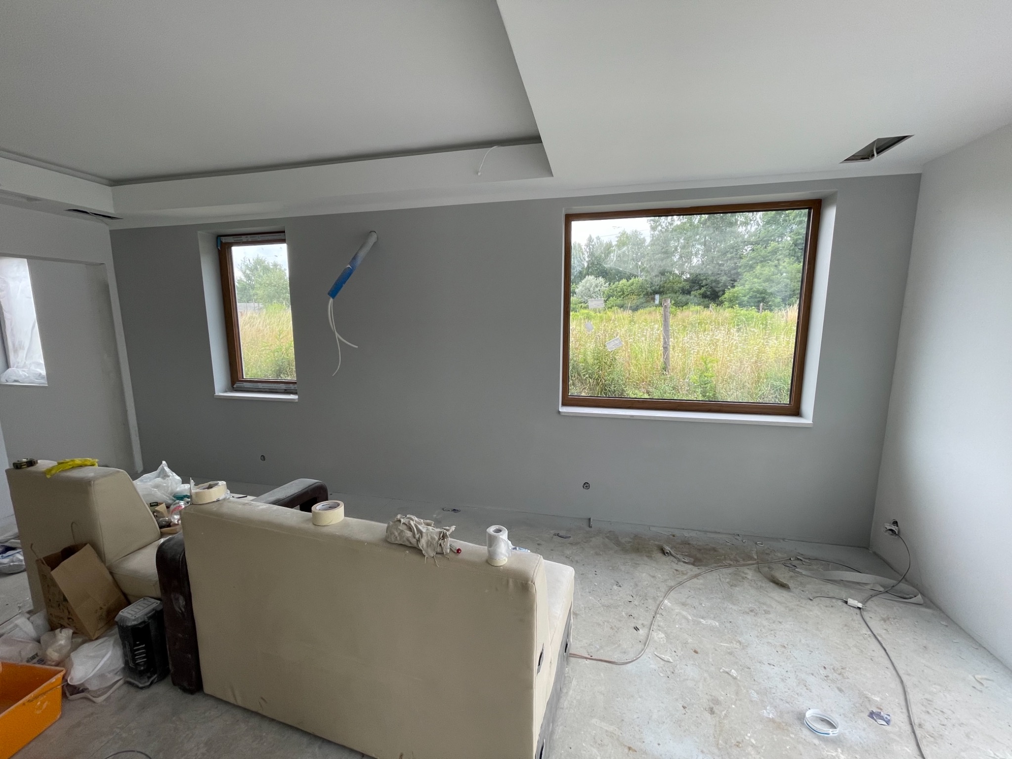 Pokój w trakcie odświeżania, ściany pomalowane na szaro, widoczne okna z drewnianą ramą, niedbale przykryte meble, pozostałości po pracach na podłodze, rura instalacyjna przytwierdzona do ściany.