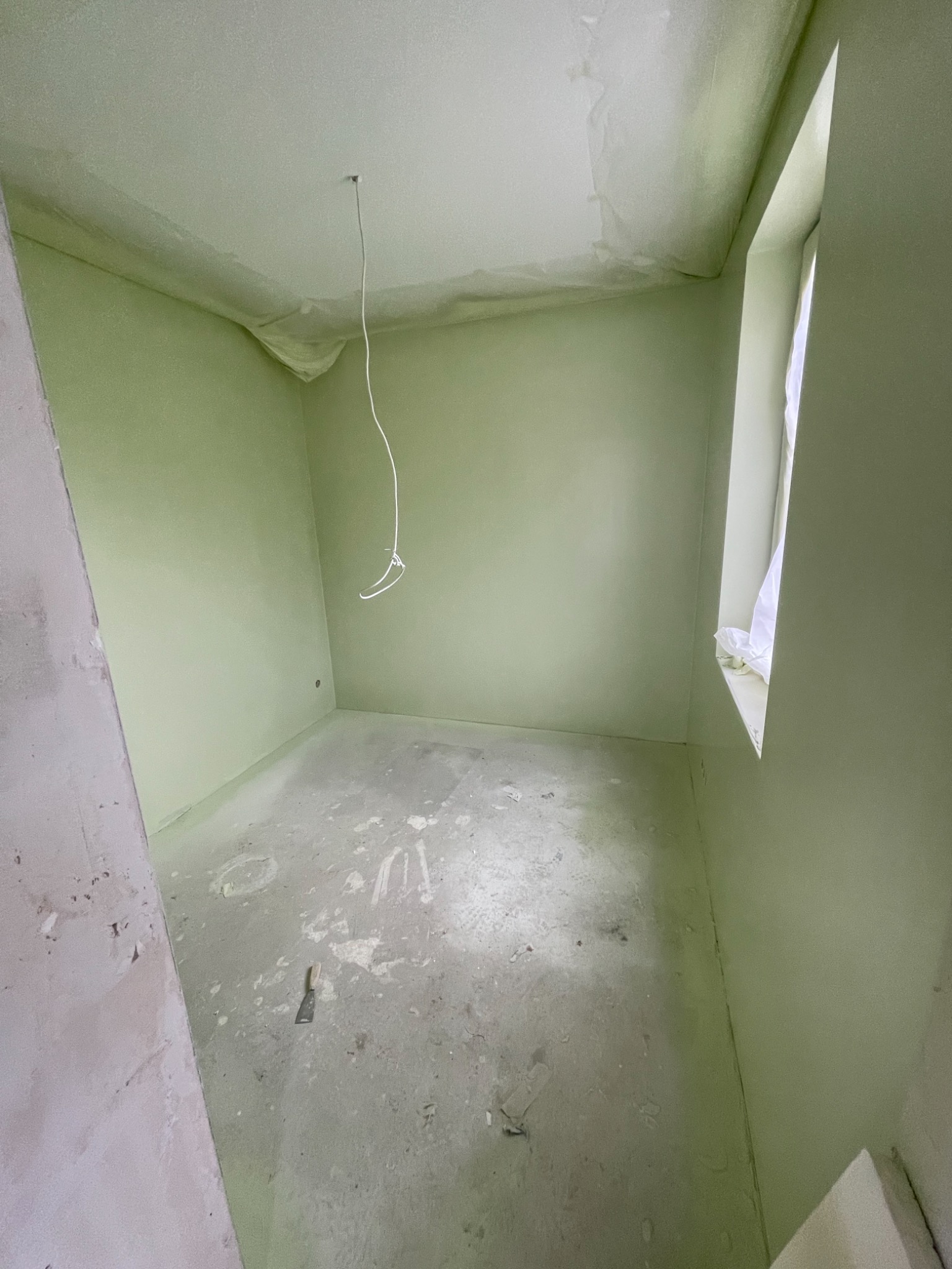 Pokój po malowaniu na zielono, z zabezpieczoną folią górną częścią ścian przy suficie i kablem zwisającym z sufitu, na podłodze widoczne ślady farby i szpachelka.