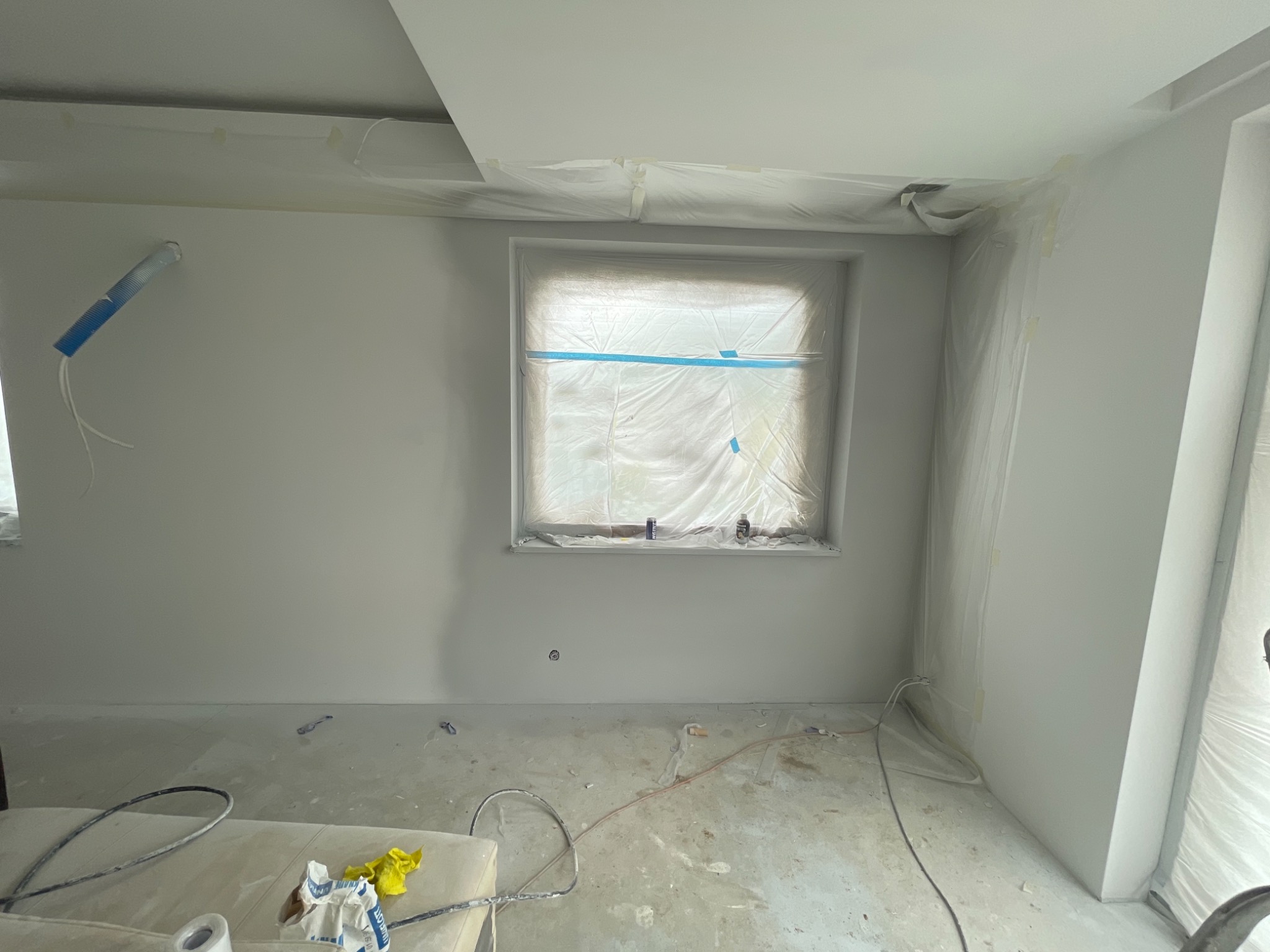 Pokój w trakcie malowania na biało, okno zabezpieczone folią, widoczne kable i akcesoria malarskie na podłodze, ściany świeżo pomalowane, sufit z widocznymi łączeniami płyt gipsowo-kartonowych.