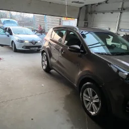 Brązowy SUV marki Kia Sportage wewnątrz myjni, z białym Renault Clio w tle i osobą wewnątrz, widoczne otwarte drzwi garażowe.