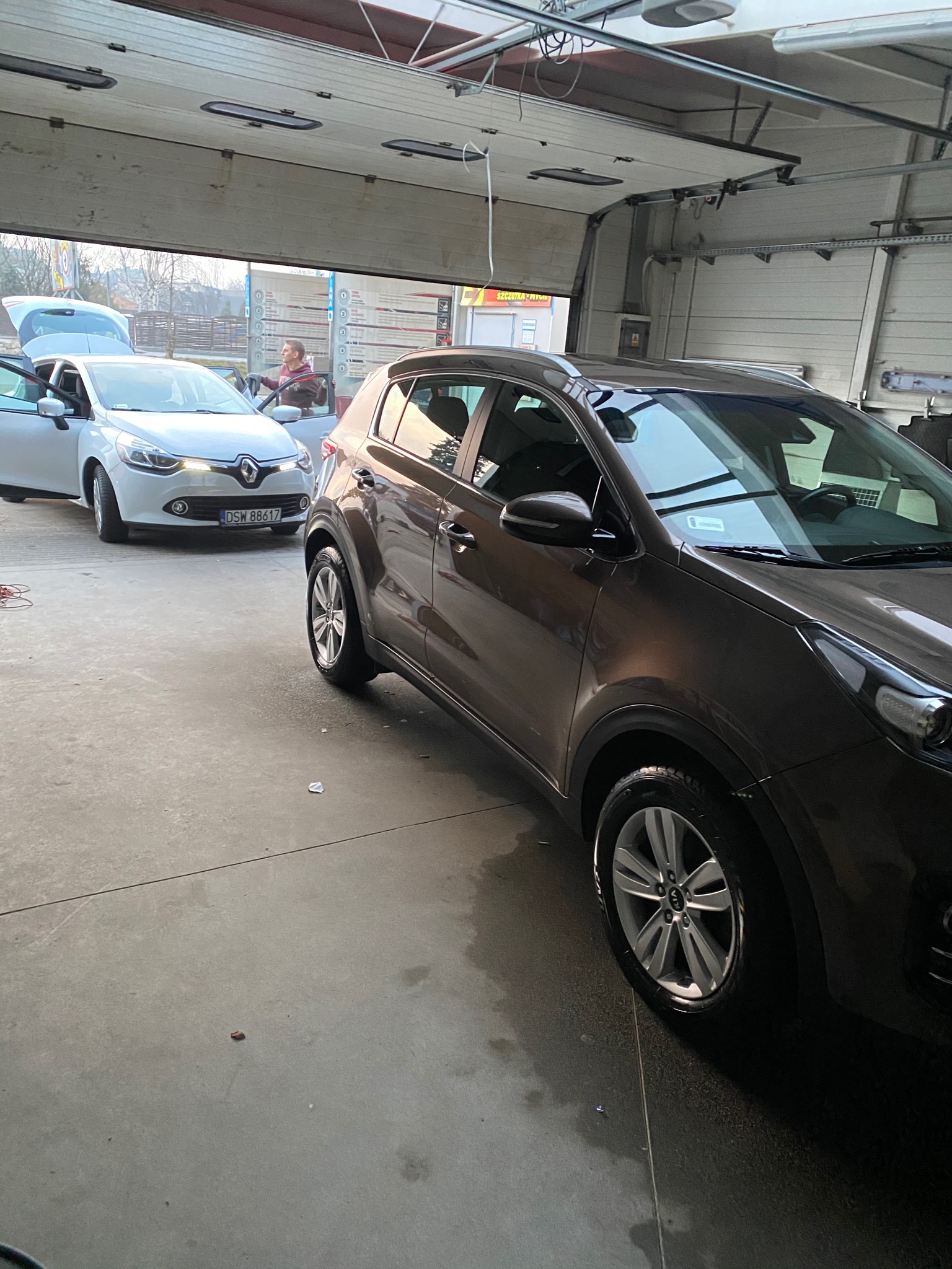 Brązowy SUV marki Kia Sportage wewnątrz myjni, z białym Renault Clio w tle i osobą wewnątrz, widoczne otwarte drzwi garażowe.