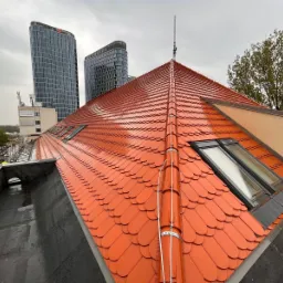 Nowy dach z czerwonej dachówki karpiówki z oknami dachowymi na tle nowoczesnych wieżowców w pochmurny dzień.