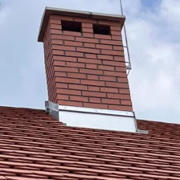 Ceglany komin z metalowym obramowaniem na dachu pokrytym czerwoną dachówką ceramiczną, widoczny fragment drabinki kominiarskiej i błękitne niebo w tle.