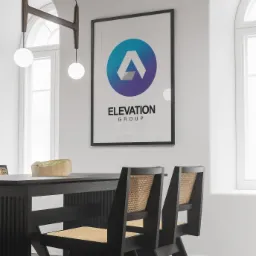 Oprawiony plakat z nowoczesnym logo Elevation Group wiszący nad czarnym stołem z owocami i krzesłami z rattanowym siedziskiem w jasnym wnętrzu.