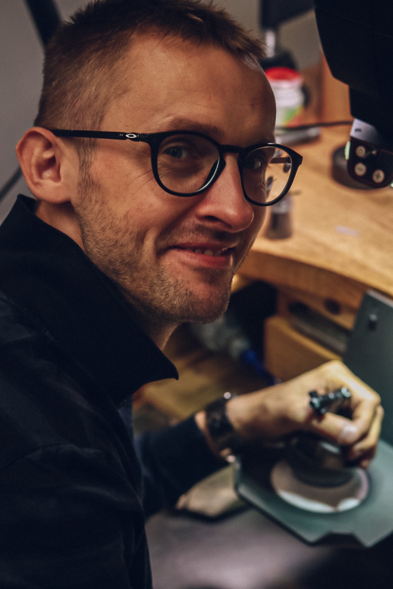 Portret uśmiechniętego mężczyzny w okularach, pracującego przy drewnianym stole z narzędziami jubilerskimi, skupionego na szlifowaniu małego elementu.