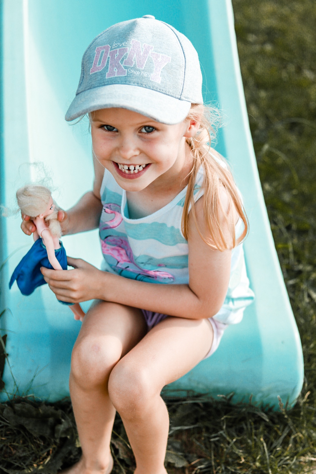 Uśmiechnięta dziewczynka w czapce DKNY siedzi na niebieskiej zjeżdżalni, trzymając lalkę z blond włosami i w niebieskiej sukience.