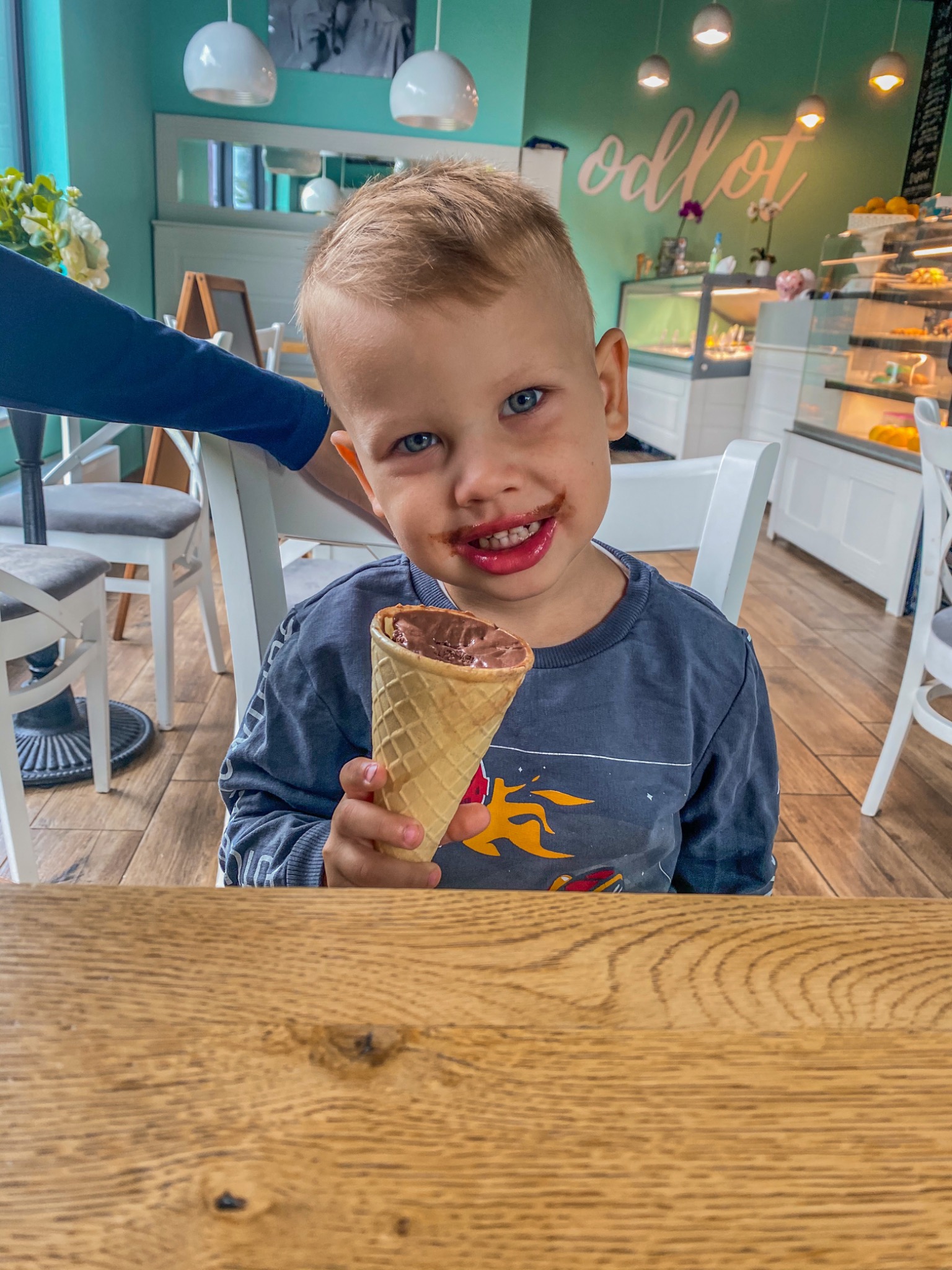 Uśmiechnięty chłopiec z niebieskimi oczami, ubrudzony czekoladowymi lodami, trzyma w ręku wafel z lodami na tle jasnego wnętrza kawiarni z drewnianym stołem na pierwszym planie.