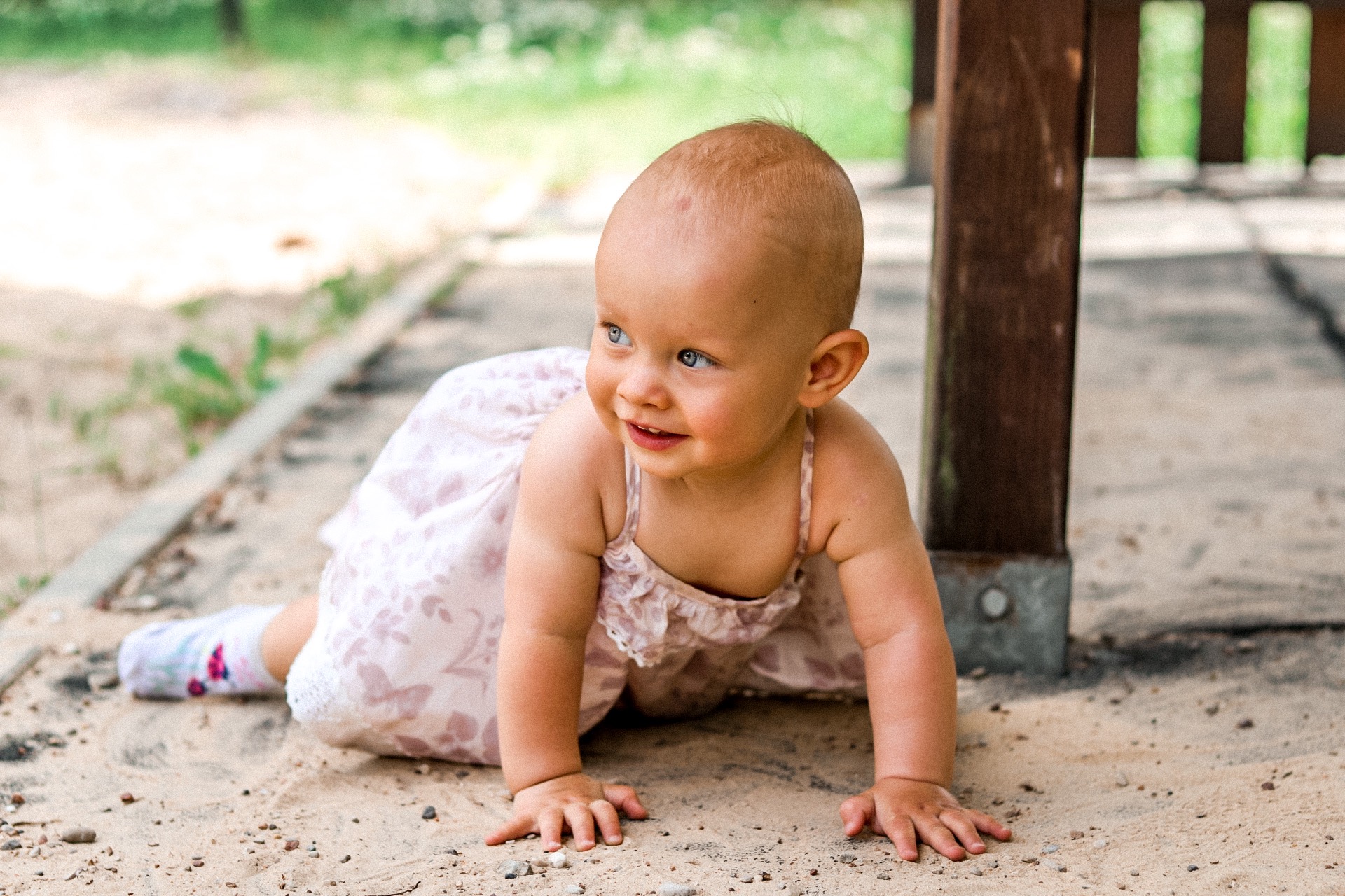 Uśmiechnięta, raczkująca dziewczynka w letniej sukience z motywem kwiatowym, w skarpetkach z aplikacją kwiatka, na piaszczystym podłożu pod drewnianą konstrukcją.