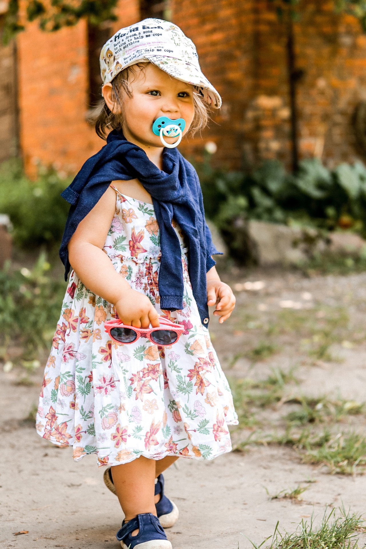 Urocza dziewczynka z blond włosami, w kwiecistej sukience i czapce z daszkiem, trzymająca różowe okulary przeciwsłoneczne i smoczek, pozująca na zewnątrz na tle ceglanego budynku.