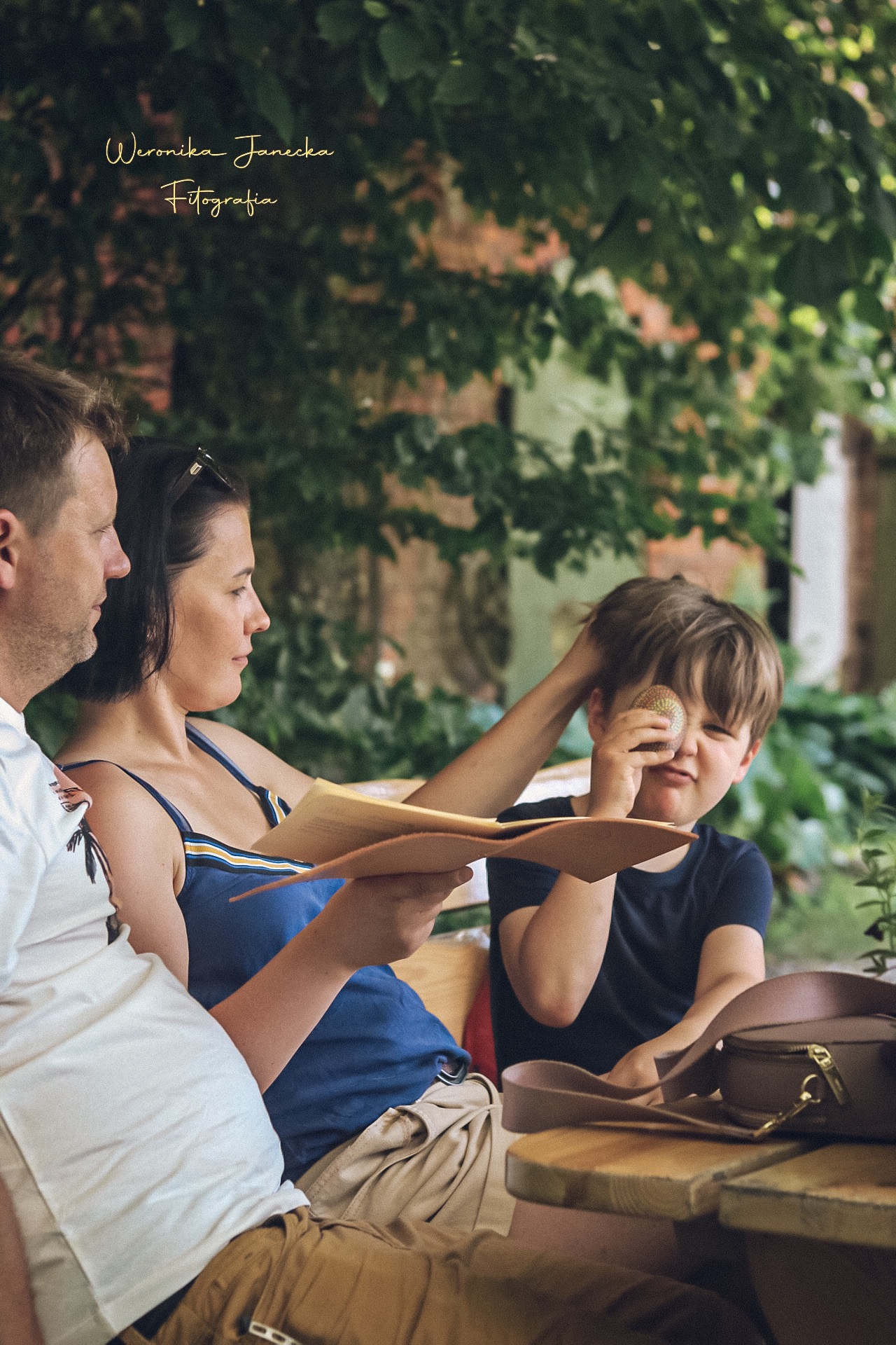 Rodzina siedząca przy drewnianym stole w plenerze, kobieta trzyma menu, a chłopiec zasłania oko małą piłeczką.