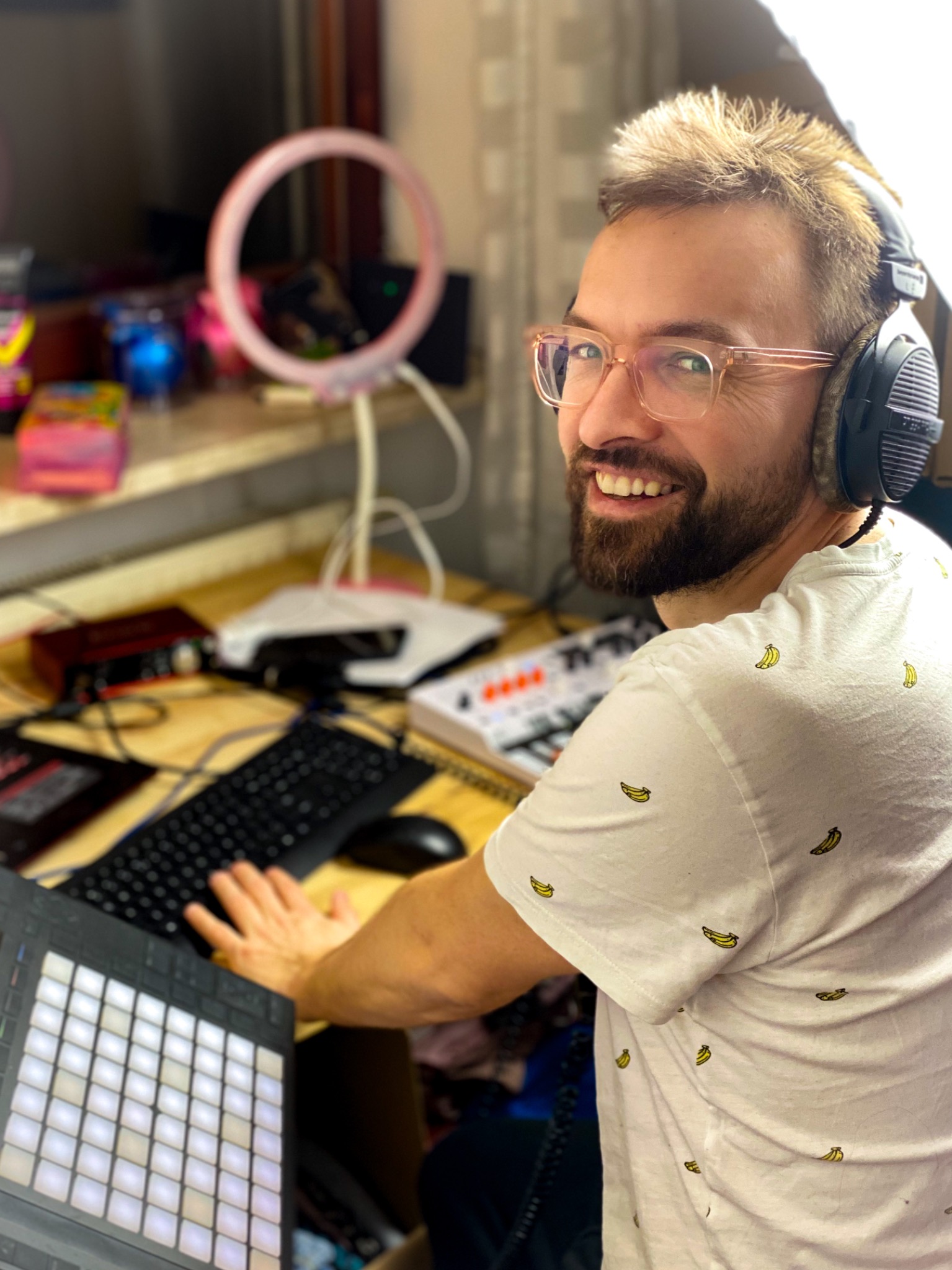 Uśmiechnięty mężczyzna w okularach i słuchawkach, siedzący przy biurku z klawiaturą, panelem kontrolnym i lampą pierścieniową, ubrany w białą koszulkę w banany.