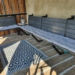 Drewniana ławka z szarym obiciem i metalowymi elementami, ustawiona na tarasie z drewnianą podłogą i białym żwirkiem, obok czarno-biały dywanik z geometrycznym wzorem, w tle drewniana ściana...