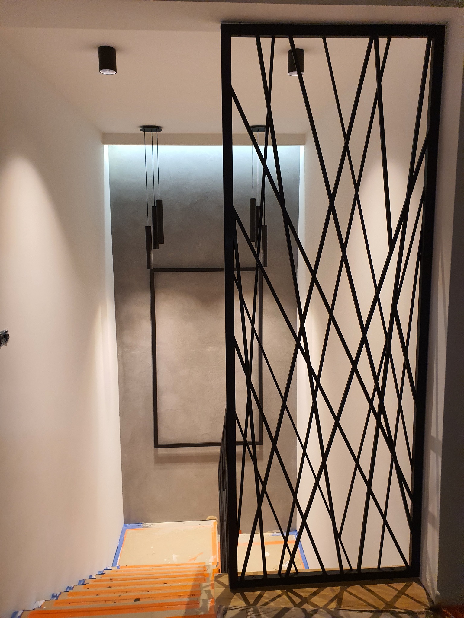 Nowoczesna, ażurowa ścianka działowa z czarnych metalowych profili w korytarzu, widoczne schody w trakcie remontu zabezpieczone folią i taśmą, minimalistyczne lampy sufitowe.