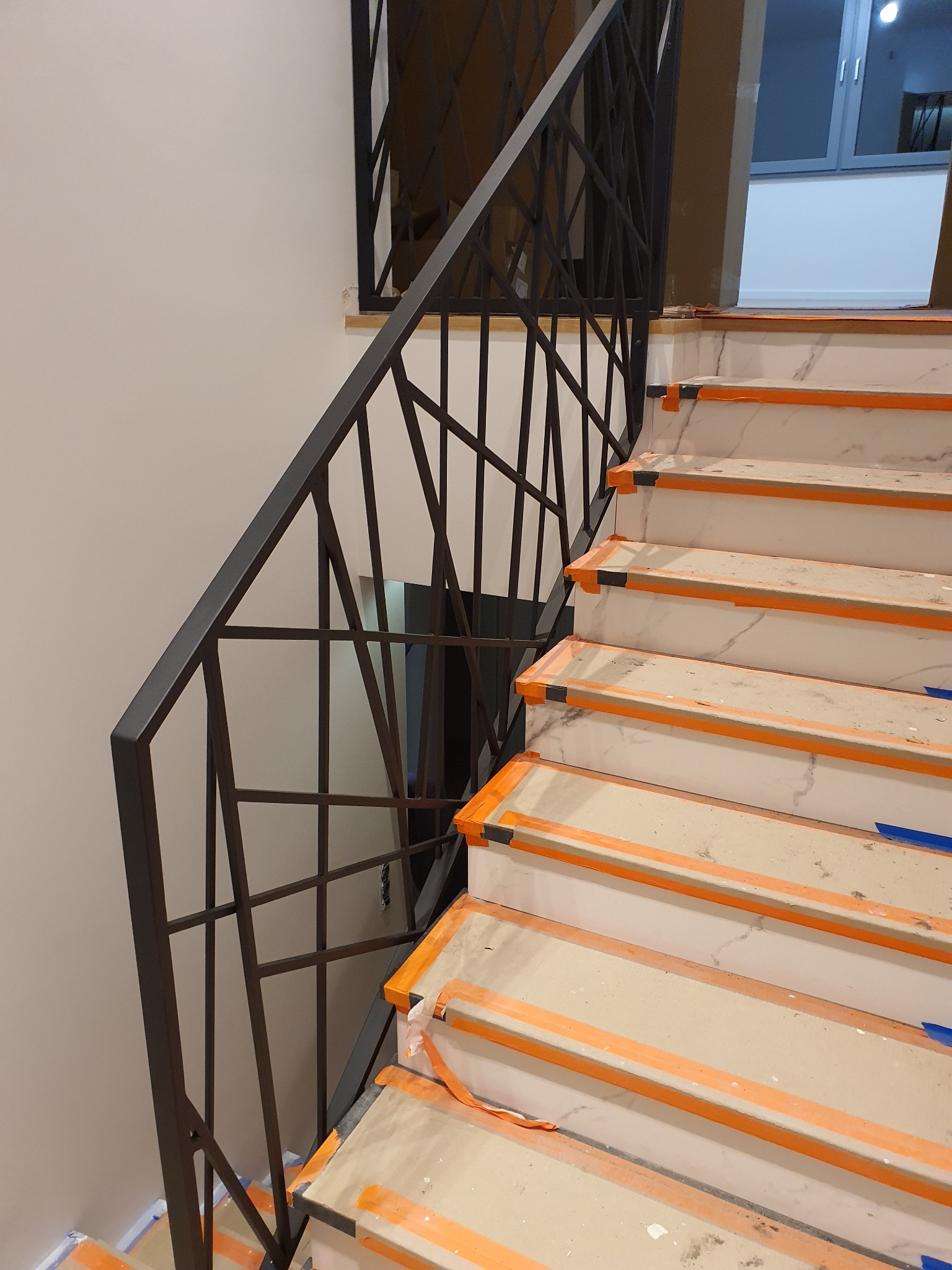 Schody z marmurowymi stopniami zabezpieczone pomarańczową taśmą malarską i nowoczesną, geometryczną balustradą z ciemnego metalu.