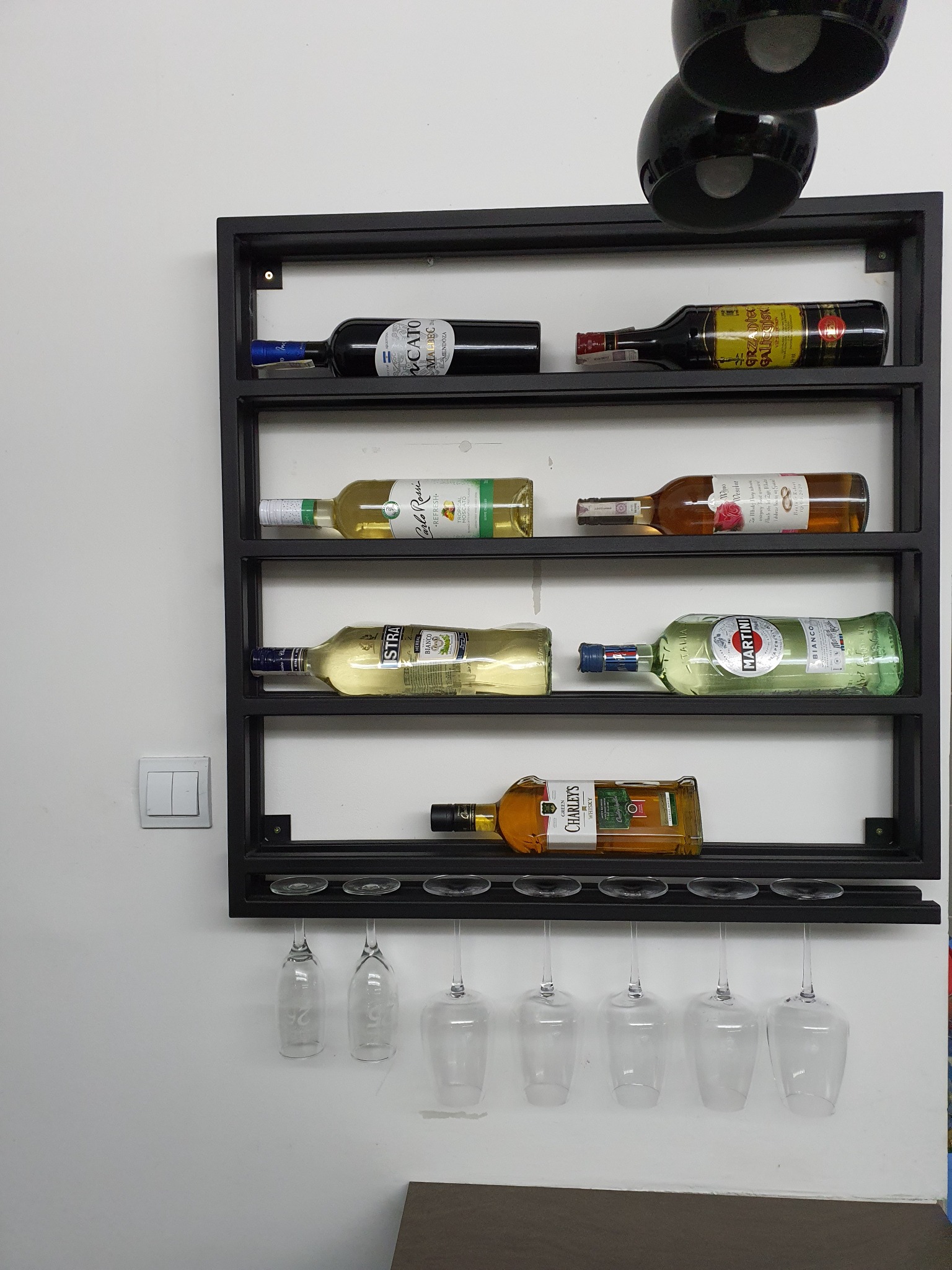 Czarna, metalowa półka na wino z uchwytami na kieliszki, zamontowana na białej ścianie w Golęczewie, eksponująca różnorodne butelki wina i whisky.