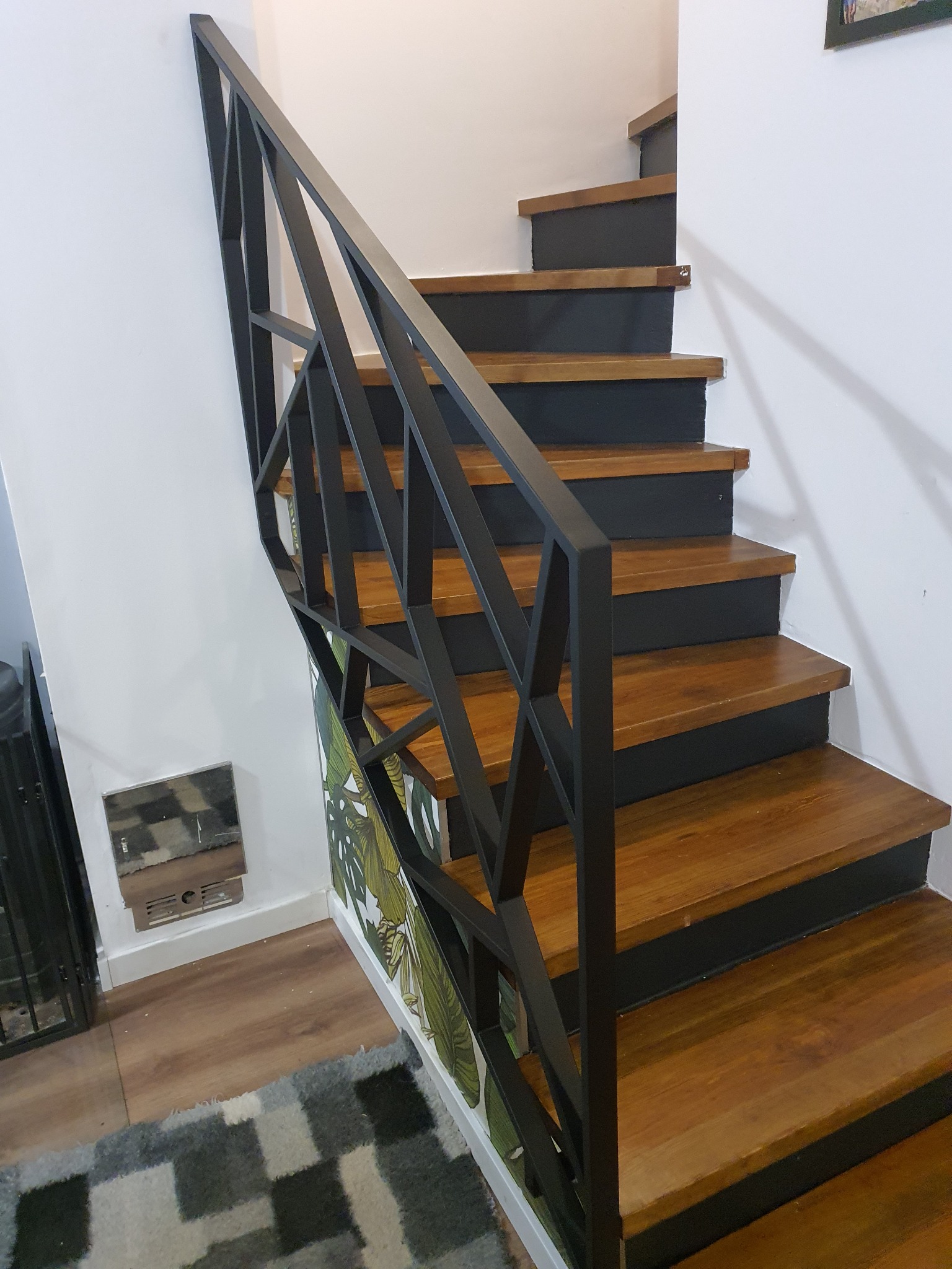 Schody drewniane z czarnymi podstopnicami i nowoczesną, geometryczną, metalową balustradą, obok ściana z motywem liści
