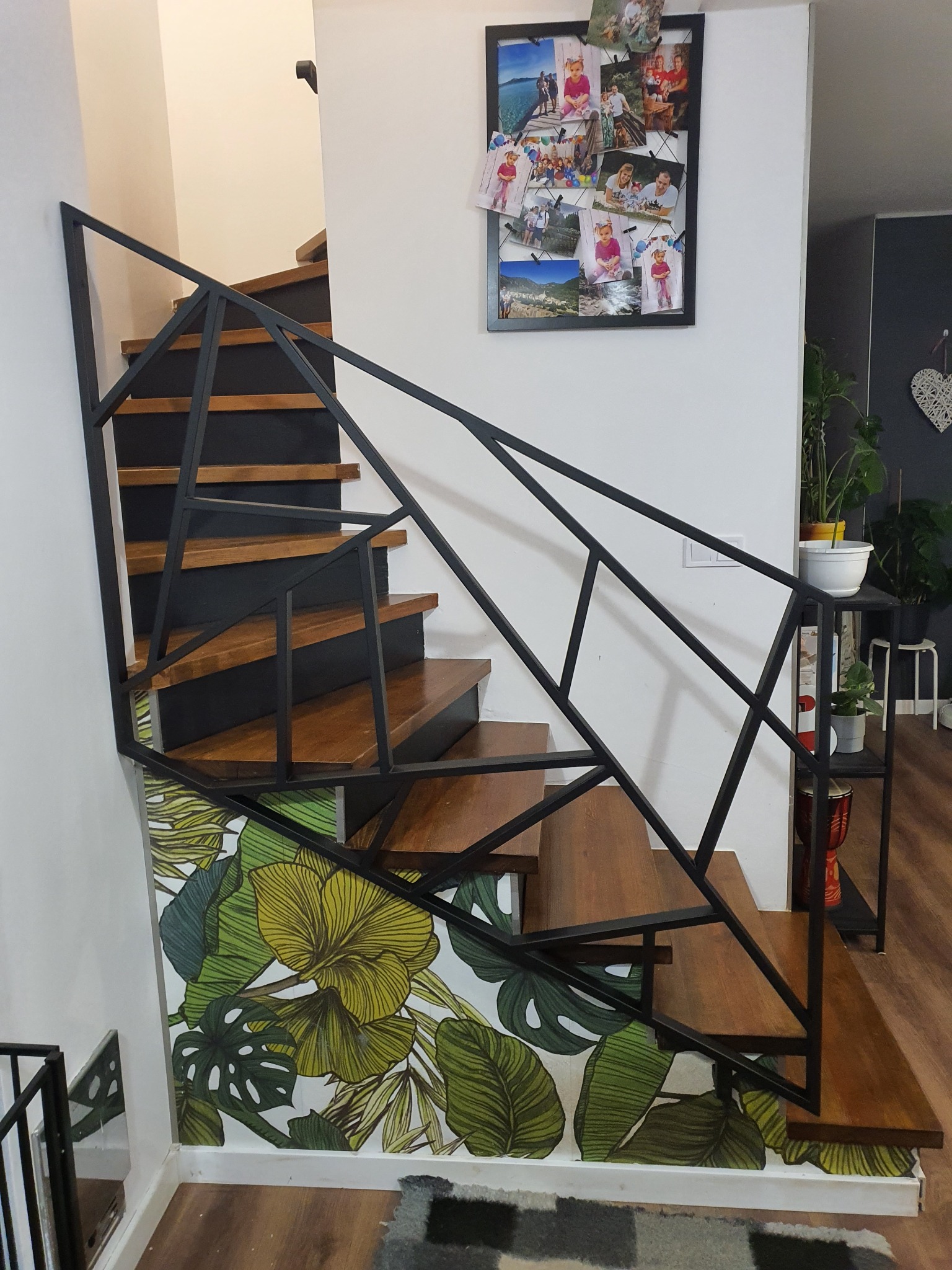 Schody drewniane z czarną, geometryczną balustradą, ozdobione tapetą z motywem tropikalnych liści, z widocznym fragmentem ściany z ramką na zdjęcia.