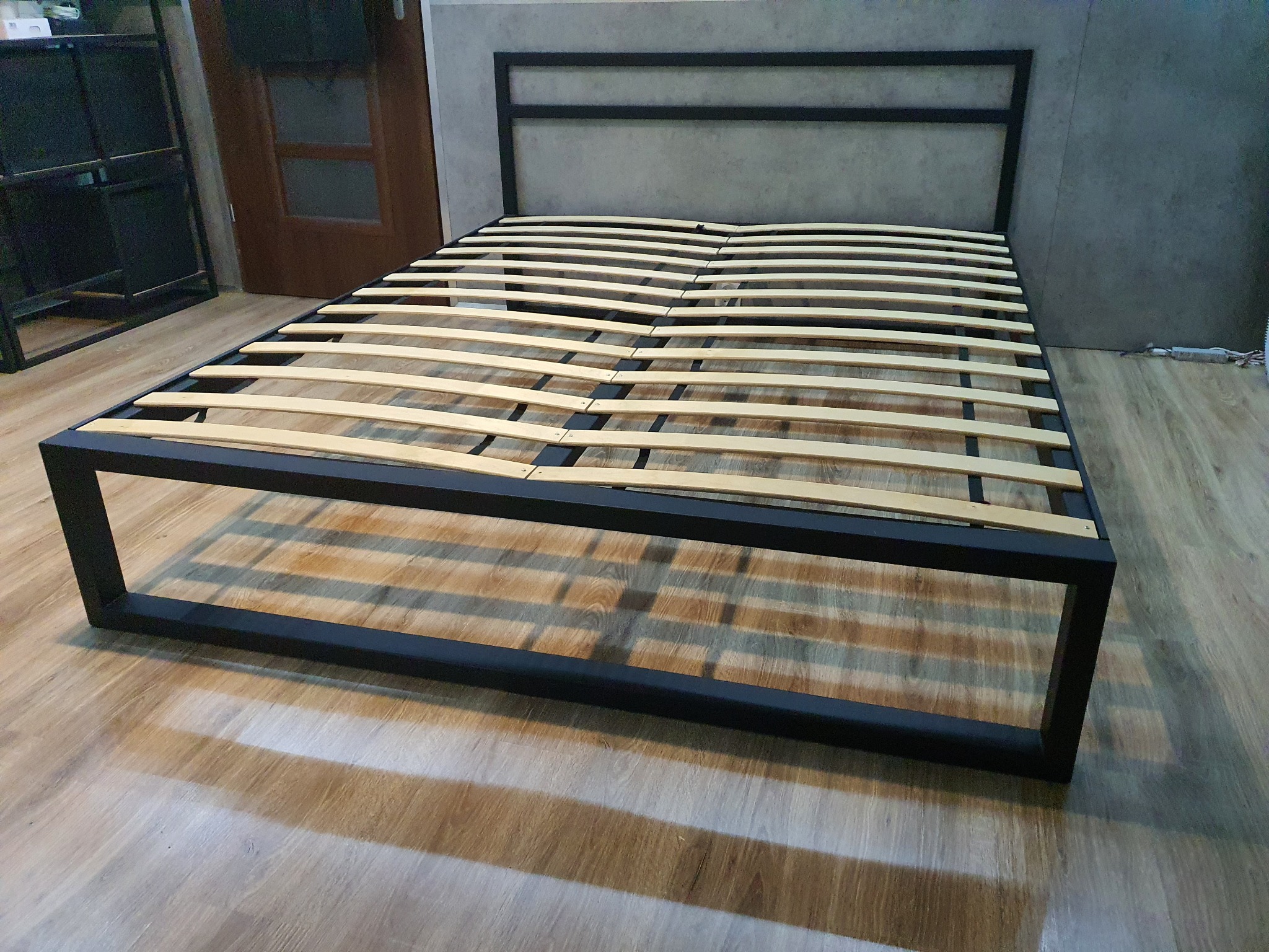 Metalowa rama łóżka z drewnianymi listwami, minimalistyczny design, surowe wnętrze, widoczna podłoga z paneli.