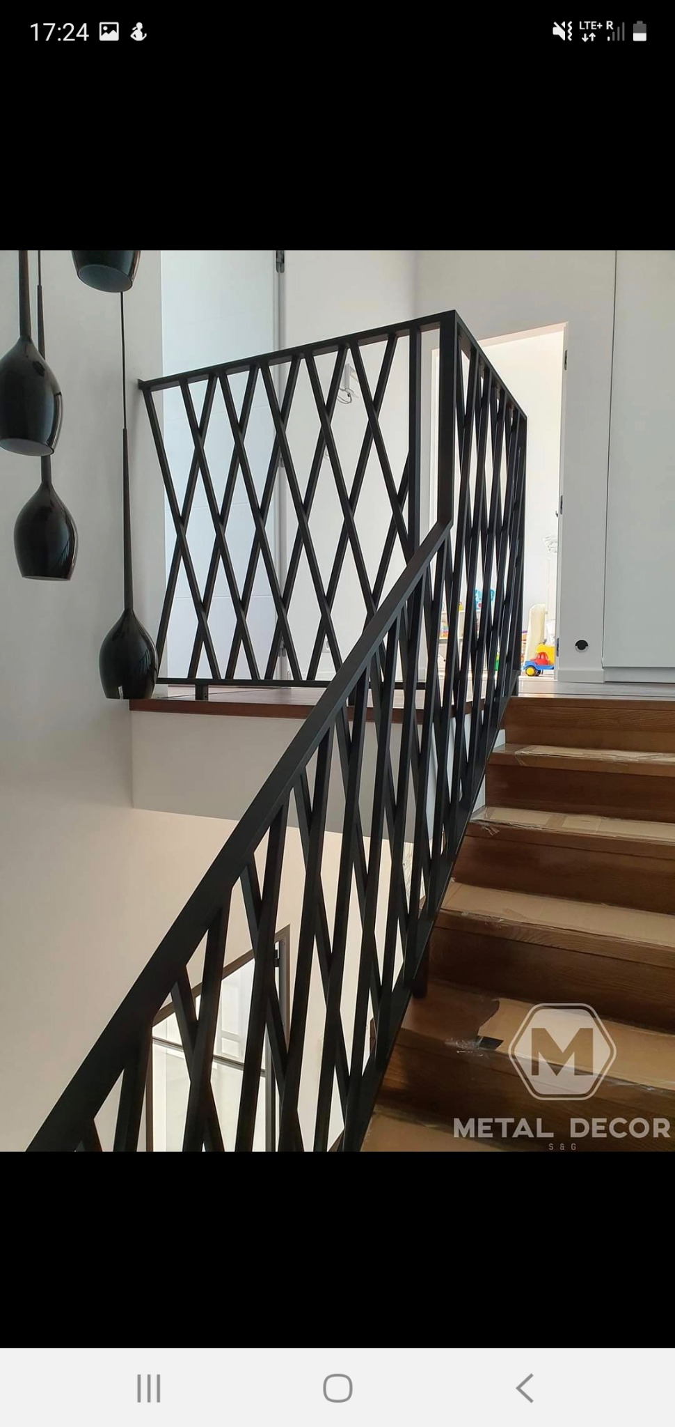 Nowoczesna balustrada schodowa z czarnych, metalowych elementów tworzących romboidalny wzór, widoczna na tle białej ściany i drewnianych schodów, z logo firmy Metal Decor w prawym dolnym rogu.