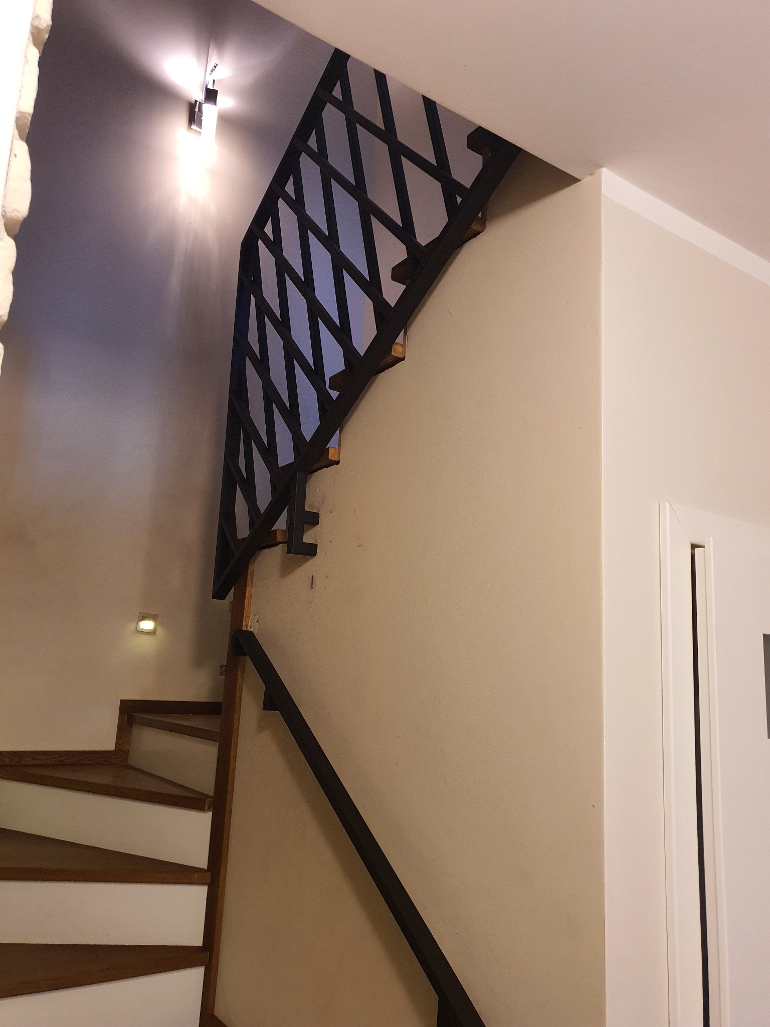 Schody drewniane z biało malowanymi podstopnicami i czarną, metalową balustradą o geometrycznym wzorze, oświetlone lampą sufitową i kinkietem.