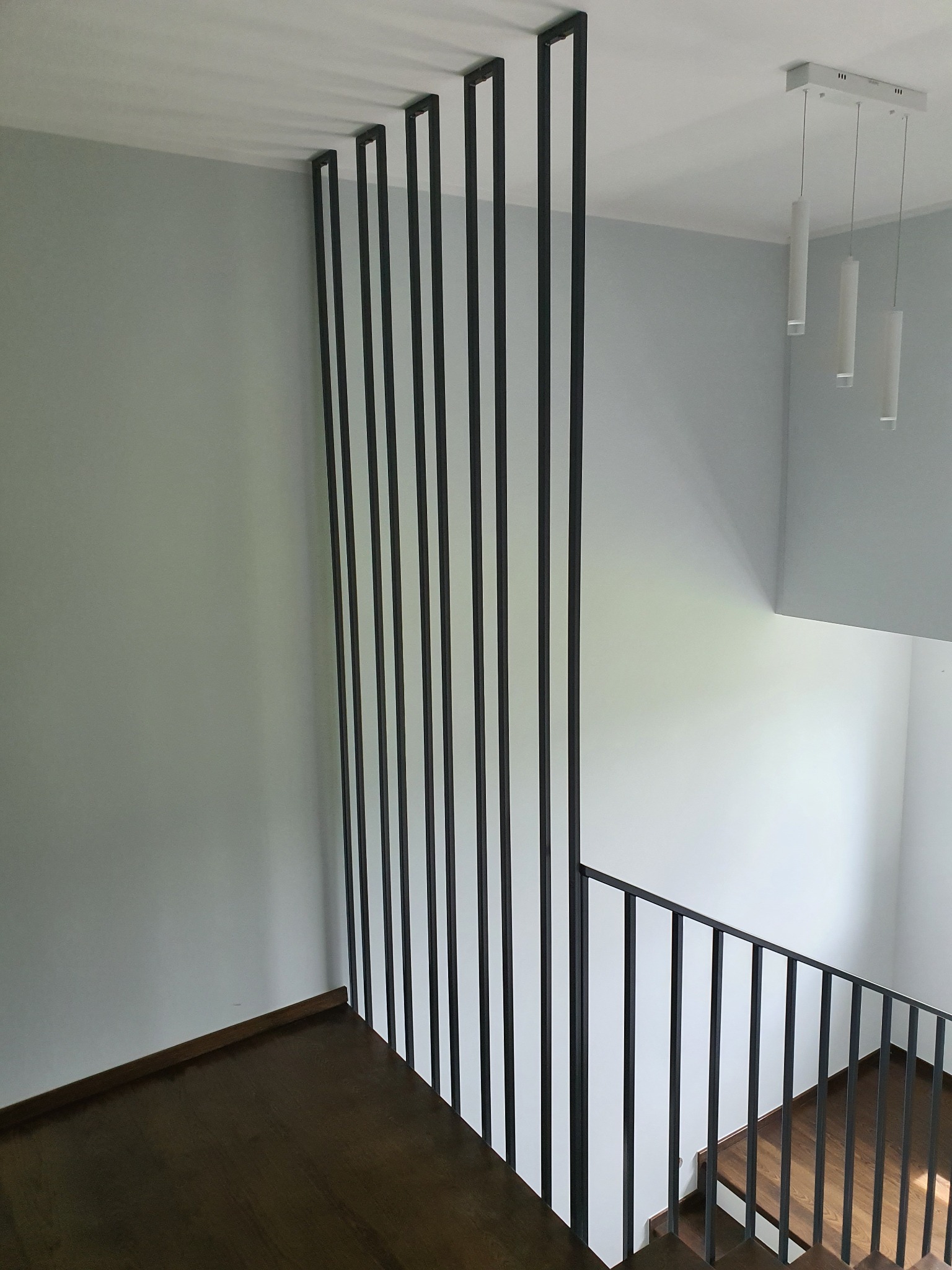 Nowoczesne schody z drewnianymi stopniami i czarną, metalową balustradą oraz pionowymi elementami dekoracyjnymi, oświetlone wiszącą lampą z trzema cylindrycznymi kloszami.
