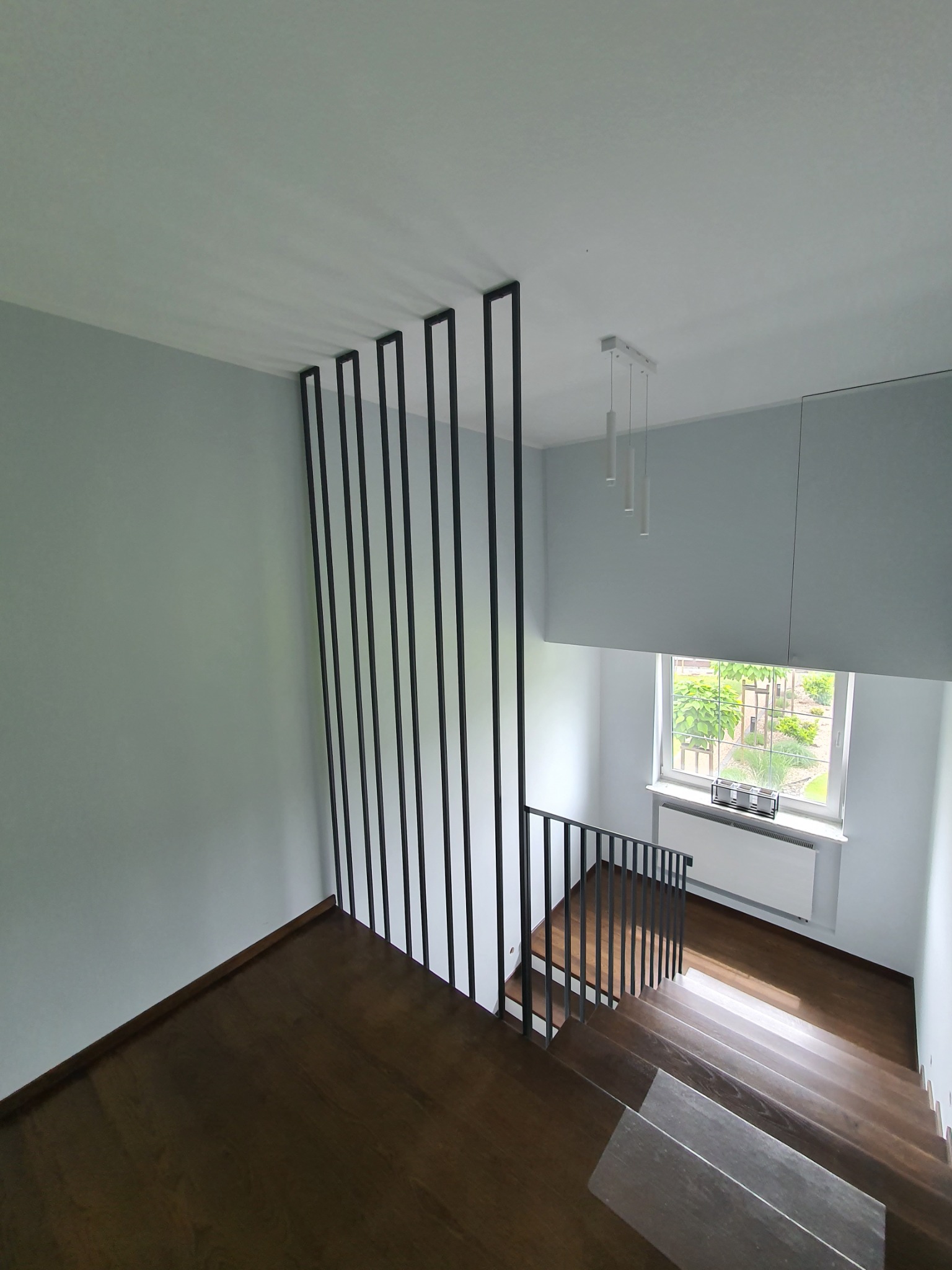 Nowoczesne schody z drewnianymi stopniami i minimalistyczną, czarną balustradą z pionowych prętów, widok z góry, jasne wnętrze z oknem i wiszącą lampą.