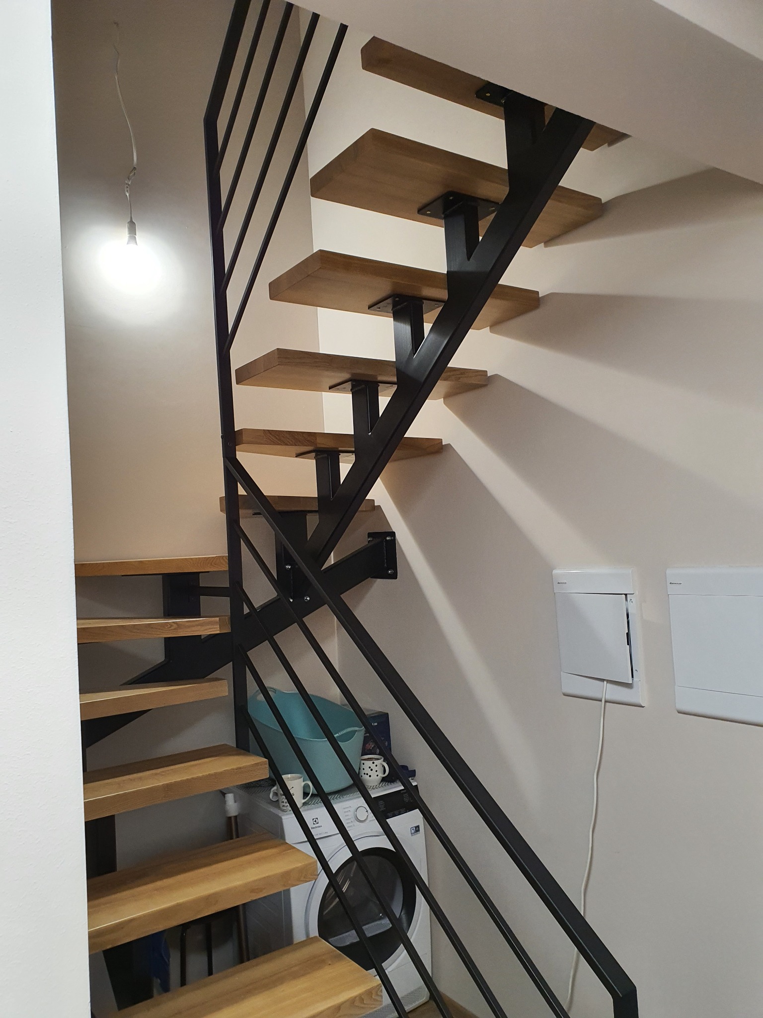 Schody drewniane z czarną metalową konstrukcją, widoczne z dołu, z pralką i koszem na pranie w tle.