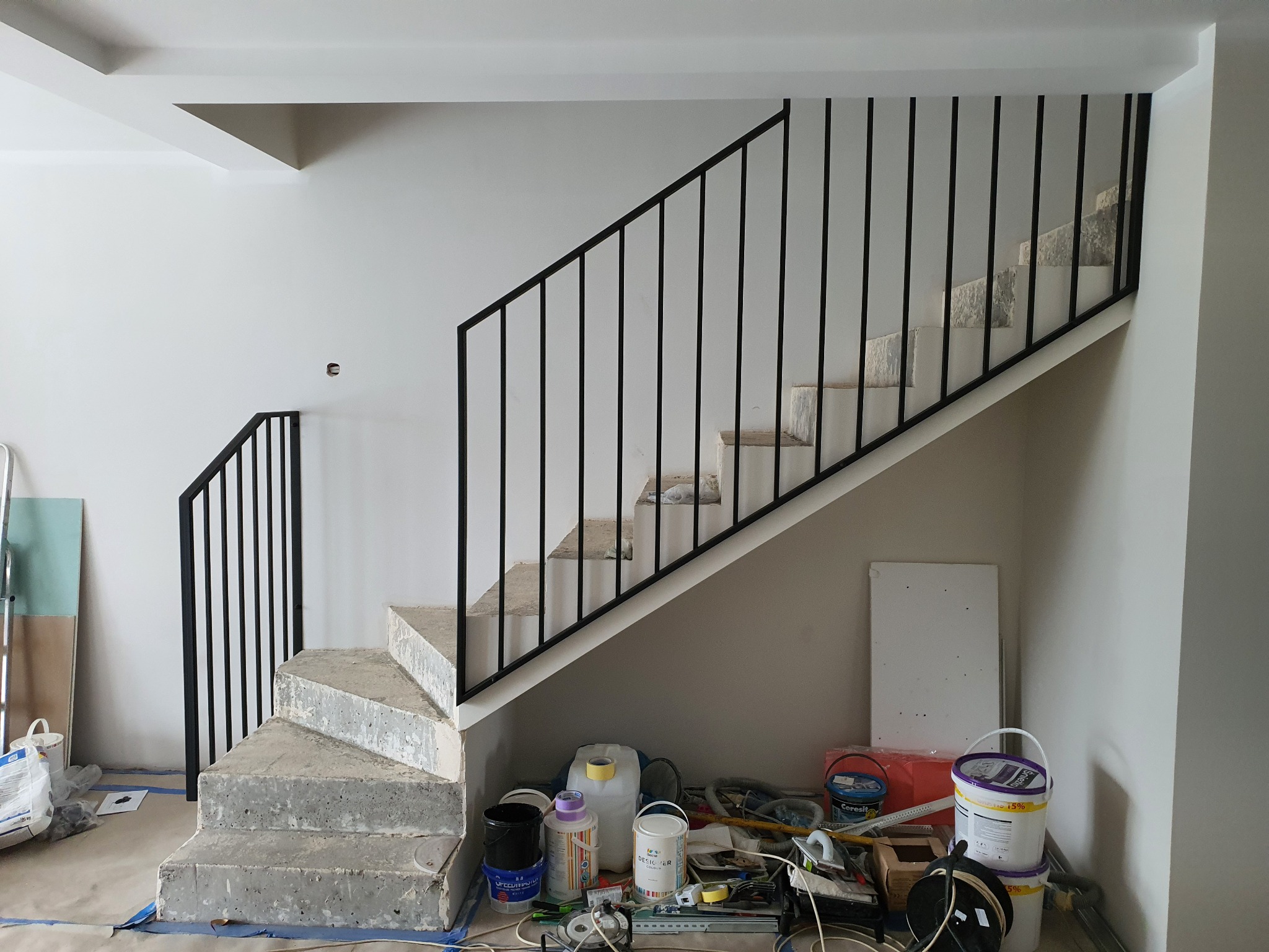 Surowe betonowe schody z czarną metalową balustradą w trakcie budowy lub remontu, widoczne narzędzia i materiały budowlane w tle, ściany w kolorze białym i szarym.
