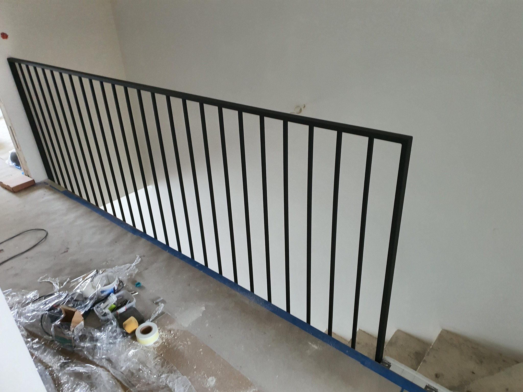 Nowoczesna, czarna balustrada schodowa z metalowymi, pionowymi prętami, zamontowana w surowym wnętrzu podczas remontu, widoczne narzędzia i folia ochronna na podłodze.