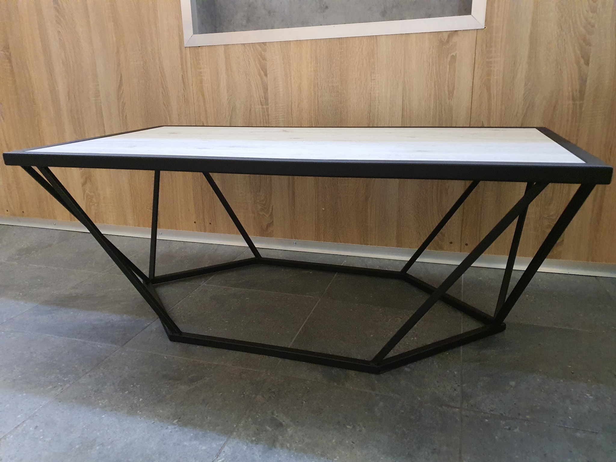 Stolik kawowy z białym blatem i geometryczną, metalową podstawą w kolorze czarnym, ustawiony na szarej podłodze, z widoczną drewnianą ścianą w tle.