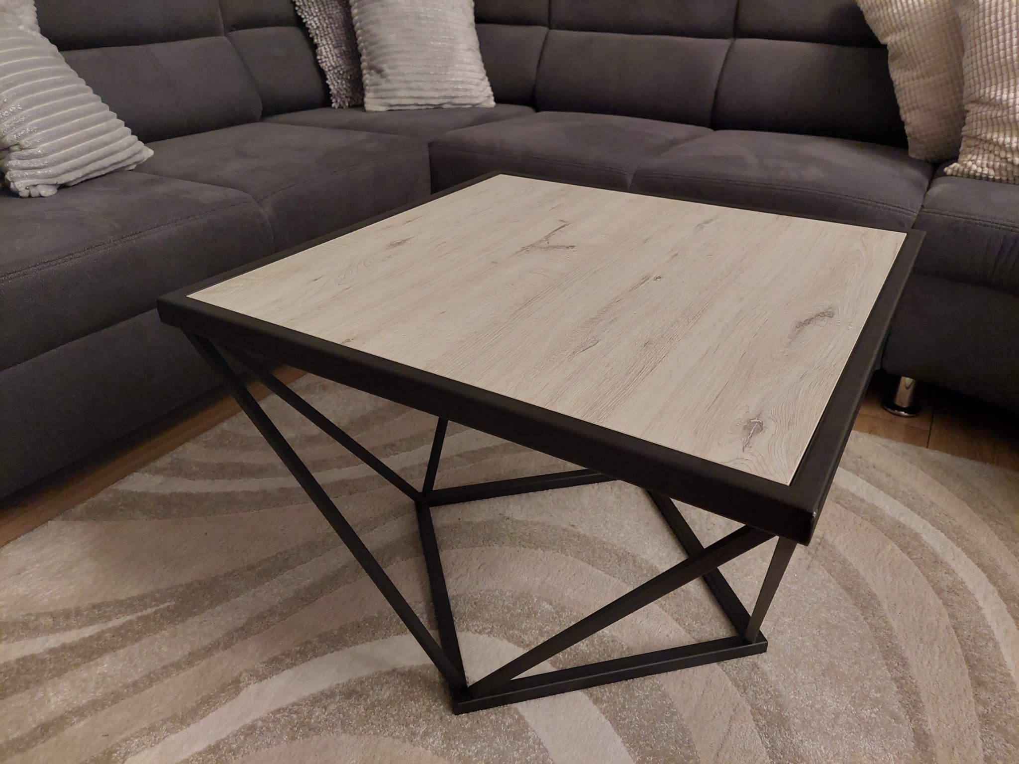 Kwadratowy stolik kawowy z blatem imitującym jasne drewno i geometryczną, metalową podstawą, ustawiony na wzorzystym dywanie, z fragmentem szarej sofy w tle.