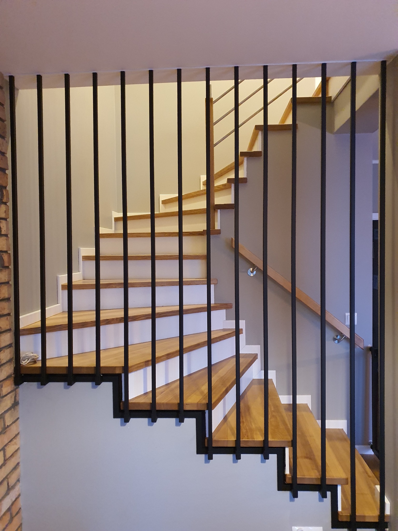 Schody drewniane z białymi podstopnicami i czarną metalową balustradą, widok przez pionowe pręty balustrady, fragment ściany z cegły po lewej stronie.