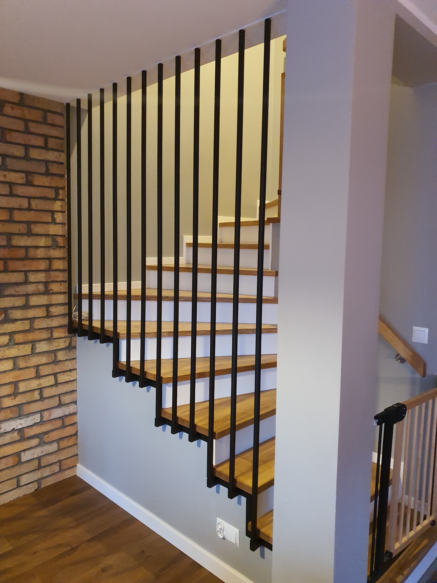 Schody drewniane z białymi podstopnicami i czarną, minimalistyczną balustradą z pionowych prętów, widoczne na tle ściany z cegły i szarej ściany w nowoczesnym wnętrzu.