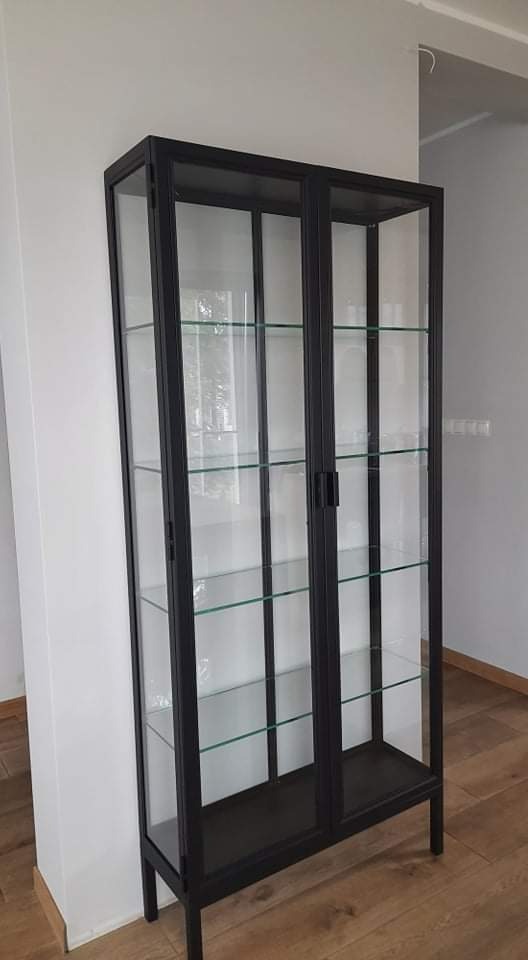 Czarna, metalowa witryna z przeszklonymi drzwiami i półkami, ustawiona w rogu pomieszczenia z drewnianą podłogą i białymi ścianami.